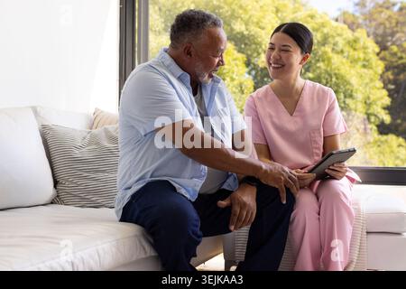 Asiatische Krankenschwester in rosafarbenen Peelings und afroamerikanische Seniorin, die auf weißem Sofa sitzt und eine Tablette überprüft Stockfoto