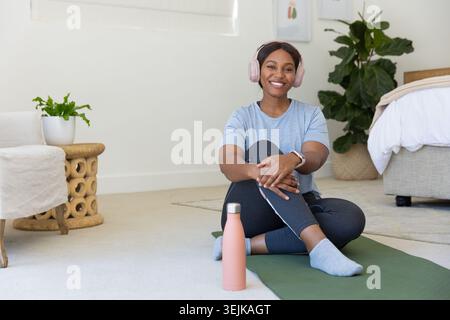 Erwachsene afrikanische Frau, die auf grüner Yogamatte im Schlafzimmer sitzt, rosa Headset trägt und eine Wasserflasche hält Stockfoto