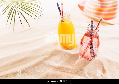 Tropische frische kalte Fruchtsäfte rosa und gelbe Säfte in nassen Glasflaschen mit Stroh, Sonnenhut, grünem Palmblatt im Sonnenlicht auf weiß Stockfoto