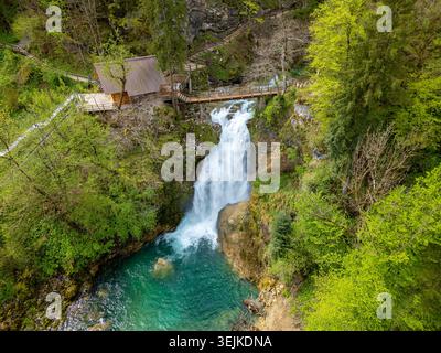 Blick aus der Vogelperspektive auf den Slap Sum Wasserfall mit einer Holzbrücke und einer kleinen Hütte umgeben von üppig grünen Wäldern in Zgornja Besnica, Kranj, Slowenien. Stockfoto