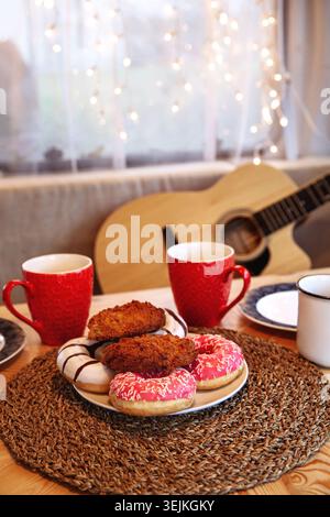 Leckere Donuts und zwei Tassen heißen Tee, Kaffee oder Kakao auf dem Tisch über der glühenden Girlande am Fenster Stockfoto