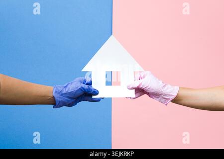 Hände in Handschuhen halten das Papierhaus auf einem farbigen Hintergrund. Schutz von Angehörigen. Prävention der Virusepidemie. Pflege des rel Stockfoto