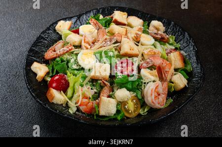 Caesar-Salat mit Tigergarnelen, Garnelensalat, Salatblättern, Wachteleiern, Kirschtomaten, parmesankäse, auf einem schwarzen Teller, Stockfoto