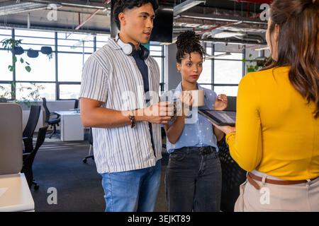 Verschiedene Kollegen stehen und diskutieren Tablets, während sie Kaffeetassen in einem hellen Büro halten. Zusammenarbeit, Teamarbeit, Präsentati Stockfoto
