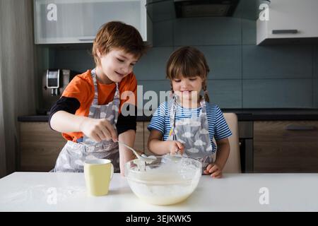Zwei Kinder kochen zusammen Teig, backen Kuchen in der Küche. Spielerische Geschwister helfen der Mutter. Stockfoto