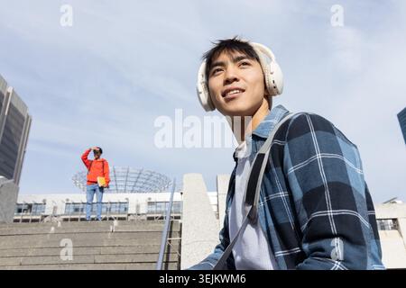 Asiatischer Mann, der auf Stufen sitzt, mit weißem Headset und Tasche, roter Jacke mit gelbem Ordner. Kopfhörer, Rucksack, Karo Stockfoto
