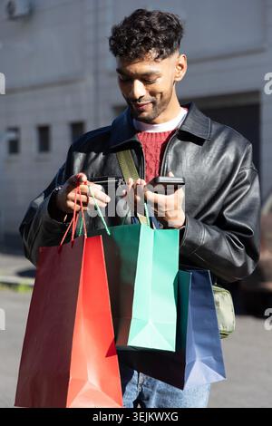 Asiatischer Mann, der in Lederjacke auf der Stadtstraße steht und 3 Einkaufstaschen hält, während er das Telefon überprüft. Urban, Shopper, Streetstyle, c Stockfoto