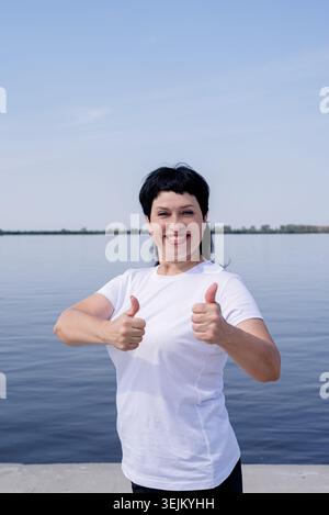 Aktive und glückliche Seniorin in Sportbekleidung, die die Daumen nach oben zeigt und am Fluss trainiert Stockfoto