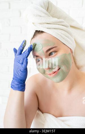 Glückliche junge kaukasische Frau in weißen Badetüchern mit Handschuhen, die eine Gesichtsmaske aus Ton aufträgt und in den Spiegel blickt Stockfoto
