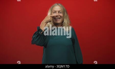 Die Seniorin zeigt Finger an Nase und lacht in grünem Kleid vor rotem Studio-Hintergrund; verspielte Freude. Stockfoto
