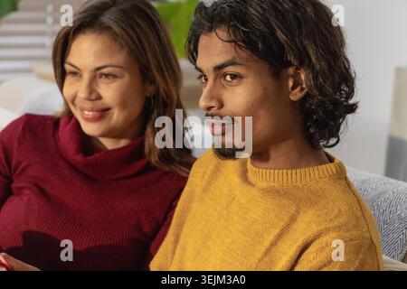 Ein Paar sitzt auf der Couch im Wohnzimmer, lächelt und trägt Strickpullover, während es ein rotes Telefon hält. Sofa, Pflanze, Innenraum, Heim, Stockfoto
