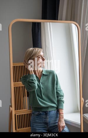Entzückte Frau, die sich glücklich und zufrieden mit der neuen Haarfarbe oder dem neuen Haarschnitt fühlt, vor dem Spiegel steht und Reflexion genießt. Smi Stockfoto