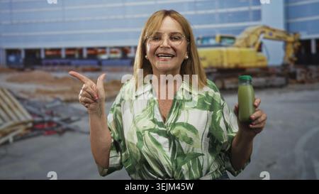 Frau, die grüne Saftflasche hält, zeigt Finger neben Baugebäude durch Bagger; Vertrauen gesunde Lebensweise. Stockfoto