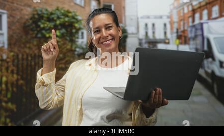 Frau, die den Laptop hält, zeigt mit dem Finger nach oben auf der Straße mit Bürgersteig und geparktem Van am Ziegelgebäude; Zuversicht Optimismus. Stockfoto