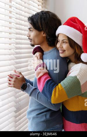 Diverse Paare umarmen sich zu Hause durch Jalousien, Frau mit Weihnachtsmütze, Pullover, Mann mit rotem Becher. Gemütlich, intim, romantisch, Dayli Stockfoto