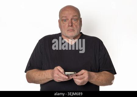 Schockierter Seniorenmann, der ein Smartphone hält und die Kamera isoliert auf weißem Hintergrund betrachtet. Stockfoto