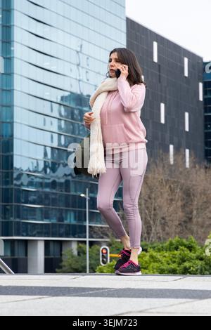 Frau in ihren dreißiger Jahren, die auf dem plaza steht und auf dem Smartphone spricht, trägt einen rosafarbenen Hoodie und hält eine Oliventasche. Fußgänger, Stadt, Schal, A Stockfoto