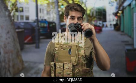 Mann Soldat in Tarnweste lächelnd und berührend einen Hundeschwanz mit der rechten Hand auf der Brust, während er auf einer Straße ein Fernglas in der Taille hält; Stolz Pflicht VI Stockfoto