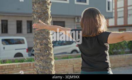 Frau, die Kopfhörer trägt, Arme gestreckt und Hände offen, tanzt auf der Straße in der Nähe von Palmen und Ziegelmauern; Freude Freiheit. Stockfoto