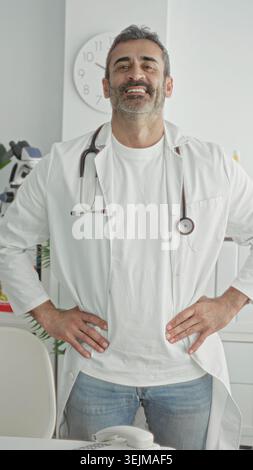 Mann Arzt mit Stethoskop an Hüften stehend in der Klinik mit Labormantel und Jeans, Uhr und Telefon sichtbar; Vertrauen. Stockfoto