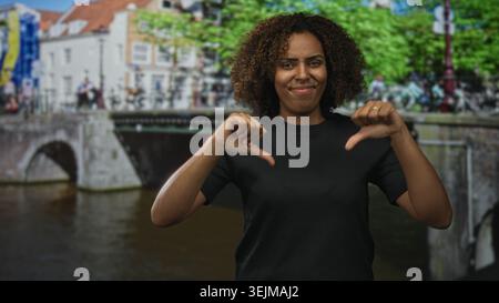 Frau zeigt auf einer amsterdamer Straße an einer Kanalbrücke mit Fahrrädern und Bäumen die Daumen nach unten; Kritik an der Missbilligung. Stockfoto