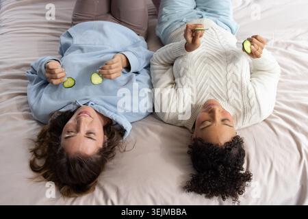Verschiedene Freundinnen, die auf dem Bett liegen, tragen einen Hoodie und einen Zopfstrick-Pullover mit Gurkenscheiben. Hautpflege, Verwöhnung, Relaxati Stockfoto