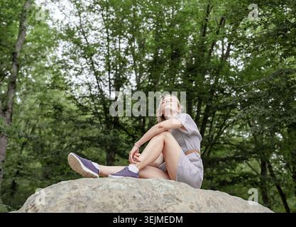 Eine junge Frau im Sommerkleid sitzt auf einem großen Stein im Wald und blickt weg Stockfoto