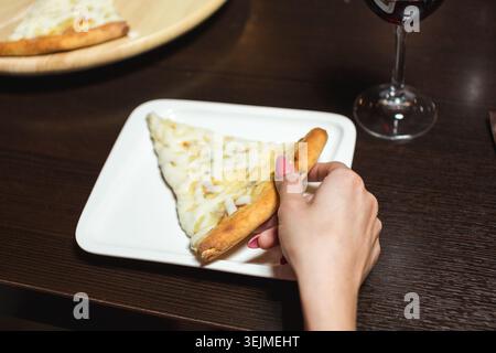 Nahaufnahme einer weiblichen Hand, die ein Stück Pizza von einem weißen Teller auf einem dunklen Tisch mit einem Glas Rotwein packt Stockfoto