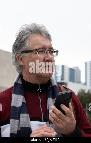 Seniorenmann, der in der Nähe der Brust auf dem städtischen plaza stand, Brille und roter Pullover trug. Rentner, Telefon, Schal, zippe Stockfoto