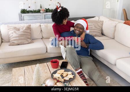 Afroamerikanisches Paar kuschelt zu Hause, Frau im Geweih, Mann im Weihnachtsmann, Kekse auf dem Tisch. Gemütlich, festlich, warm, romantisch, Stockfoto