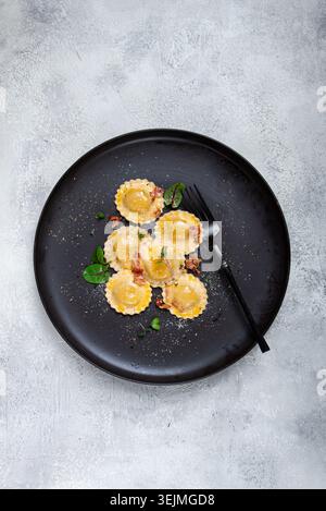 Frische Ravioli, mit Butter, Gewürzen und Kräutern, gebratener Speck, auf einem schwarzen Teller, Draufsicht, dunkler Hintergrund, keine Leute, Stockfoto