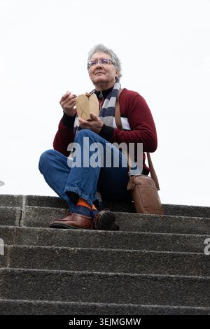 Senior Mann sitzt auf Betontreppen unter bedecktem Himmel und hält Take-out und isst mit Essstäbchen. Ältere Menschen, im Freien, Treppenhaus, Stockfoto