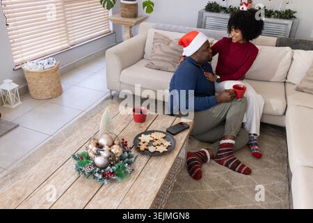 Afroamerikanisches Paar, das auf einem Sofa im Wohnzimmer sitzt und Kekse und rote Tassen teilt. Gemütlich, festlich, Urlaub, Dekor, ce Stockfoto