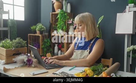 Blonde Floristin mit Schürze neben Laptop und Blumenstrauß in einem Floristikstudio; fokussierte Planung. Stockfoto