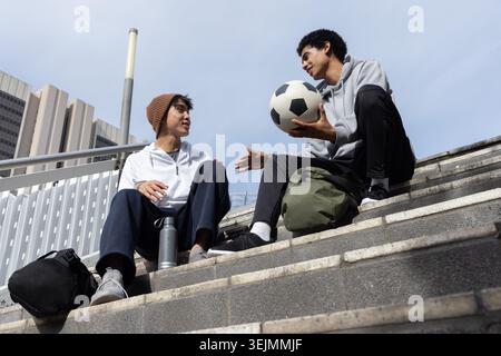 Verschiedene männliche Teenager sitzen auf Betontreppen und tragen Hoodies, die Fußball und Rucksäcke halten. Jugend, Stadt, Treppen, Freizeit, fr Stockfoto