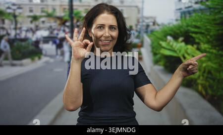 Eine Frau mittleren Alters macht mit der linken Hand ein OK-Schild und zeigt mit dem rechten Finger auf die belebte Stadtstraße; eine Geste zur Vertrauensbestätigung. Stockfoto