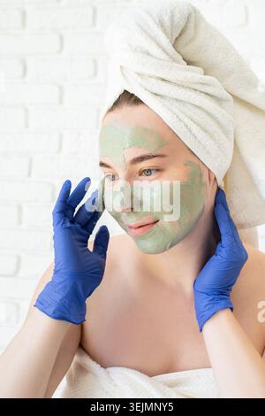 Glückliche junge kaukasische Frau in weißen Badetüchern mit Handschuhen, die eine Gesichtsmaske aus Ton aufträgt und in den Spiegel blickt Stockfoto