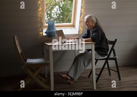 Eine ältere Frau, die von zu Hause aus mit dem Internet verbunden ist. Die entzückende ältere Frau, die einen Laptop benutzt, in einem stilvollen Haus Stockfoto