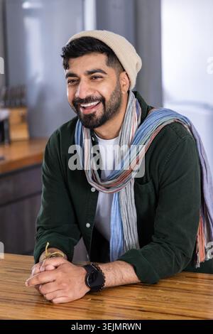 Indischer Mann lehnt sich auf Holztheke in der Küche und lächelt, während er einen Mütze-Schal und Smartwatch trägt. Porträt, Bartop, gemütlich, warm, Stockfoto