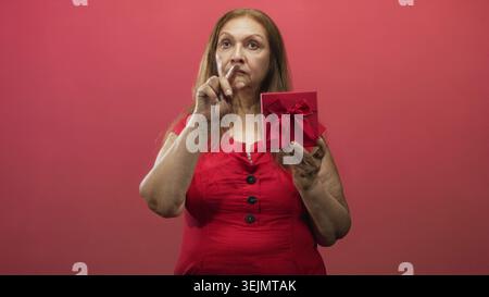 Die Frau hält eine rote Geschenkbox mit Bogen und zeigt den Zeigefinger nach oben in der Kamera, die dem Studio zugewandt ist; ernsthafte Vorfreude. Stockfoto