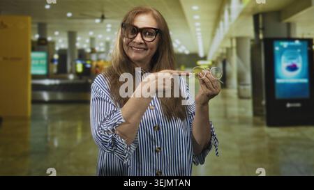 Eine Seniorin zeigt mit dem Finger auf eine Handbrille, während sie lächelt und ein Ersatzpaar in einem Flughafenterminal hält; Selbstvertrauen demonstriert. Stockfoto