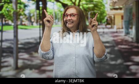 Frau mit roter Brille zeigt die Zeigefinger nach oben auf eine von einem sonnigen Baum gesäumte Straße, lächelnd und einnehmend; Freude am Optimismus. Stockfoto