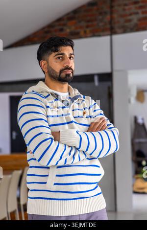 Indischer Mann, der die Arme im Loft überkreuzt trägt, einen cremegestrickten Hoodie mit Kordelzügen, einen Holztisch. Pullover, stehend, innen, mi Stockfoto
