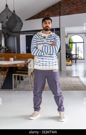 Indischer Mann mit gestreiftem Hoodie und Cargo-Hosen, stehend im Wohnbereich mit Hängelampen. Minimalistisch, modern, openpla Stockfoto
