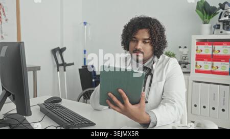Der Mann hält Tablette mit der rechten Hand und benutzt Maus mit der linken Hand, Stethoskop am Hals, im medizinischen Gebäude; fokussierte Pflege. Stockfoto