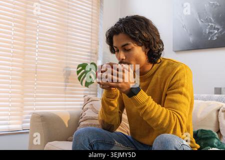 Asiatischer Mann, der Senfpullover trägt, sitzt auf Sofa im Wohnzimmer und hält rote Becher Smartwatch sichtbar. Gemütlich, minimalistisch, warm, Int. Stockfoto