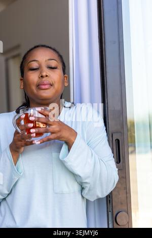 Afroamerikanerin mit hellblauem Pyjama, die an der Schiebetür steht und einen Glasbecher hält. Ruhig, gemütlich, Loungewear, Lifesty Stockfoto
