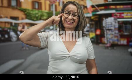 Die hispanische Seniorin zeigt mit dem Finger auf den Tempel mit Brille und weißem Oberteil auf dem geschäftigen Straßenmarkt; nachdenkliche Reflexion. Stockfoto