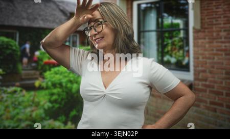 Hispanische Seniorin mit grauem Haar und Brille lächelt, Hände auf Hüften, hebt dann Hand an Stirn, um die Augen zu schattigen, während sie vor einem Gebäude steht; Co Stockfoto