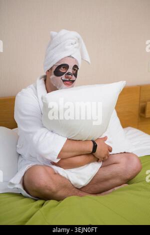 Junger Mann im weißen Morgenmantel, der mit Gesichtsmaske entspannt, junger Mann im weißen Morgenmantel, der mit Gesichtsmaske entspannt Stockfoto